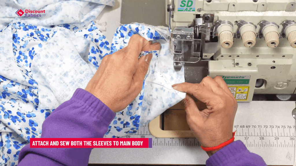 Person sewing fabric with a blue floral pattern using a sewing machine. They are attaching sleeves to a garment. A red bracelet adorns their wrist. Text on image: