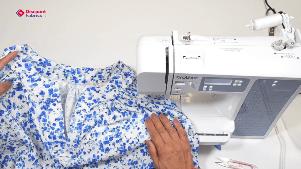 Person sewing blue floral fabric with a Brother sewing machine, following a sewing blouse tutorial. Hand adjusting fabric near the needle, scissors and thread nearby. Discount Fabrics logo in the top left corner.