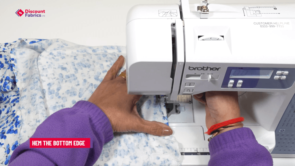 A person wearing a purple sweater uses a Brother sewing machine to hem the bottom edge of a light blue patterned fabric. A text overlay in the corner reads,