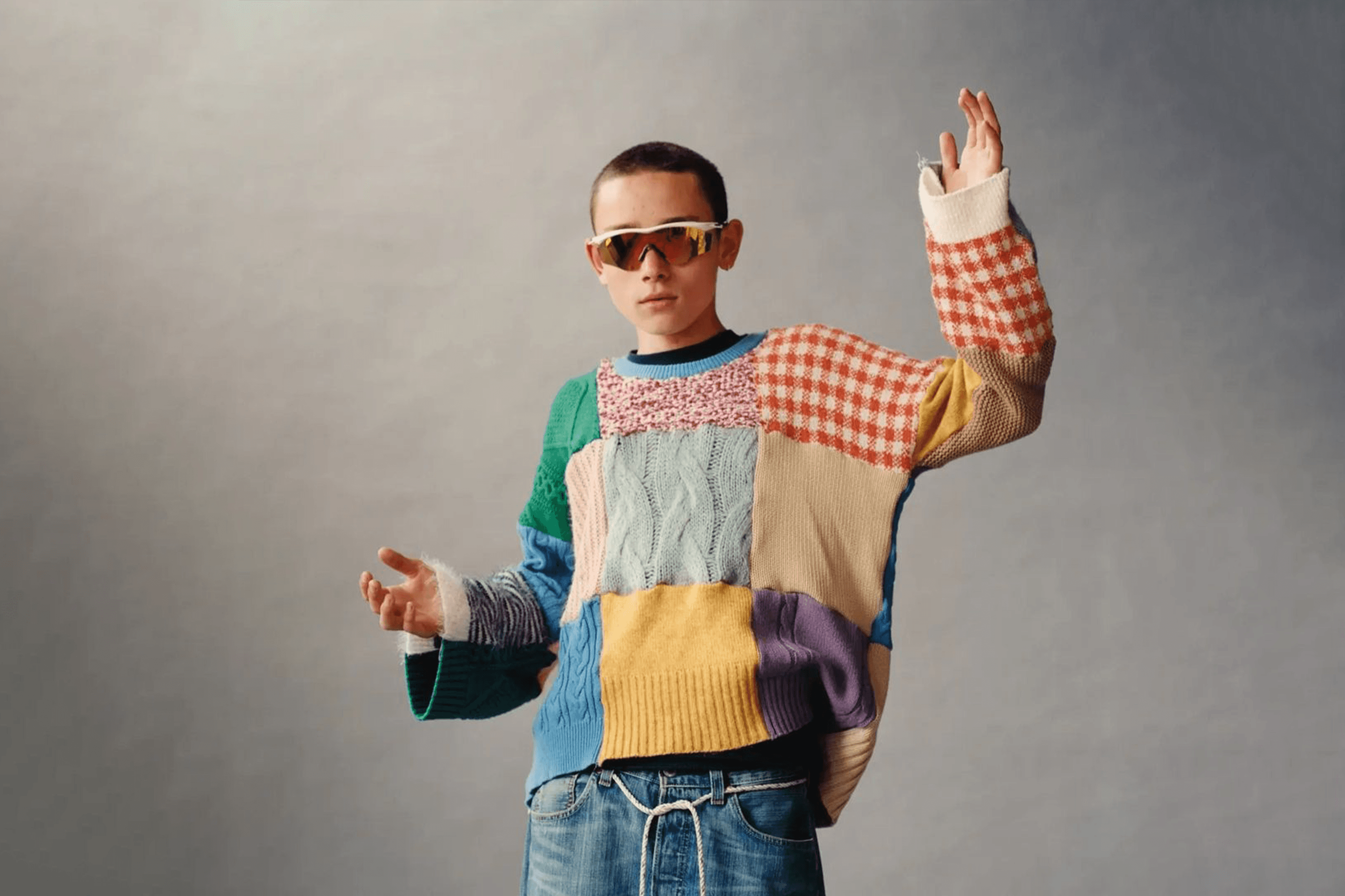 A person with short hair wears a colorful patchwork sweater, blue jeans, and reflective sunglasses. They have raised arms and stand against a gray background, striking a casual pose.