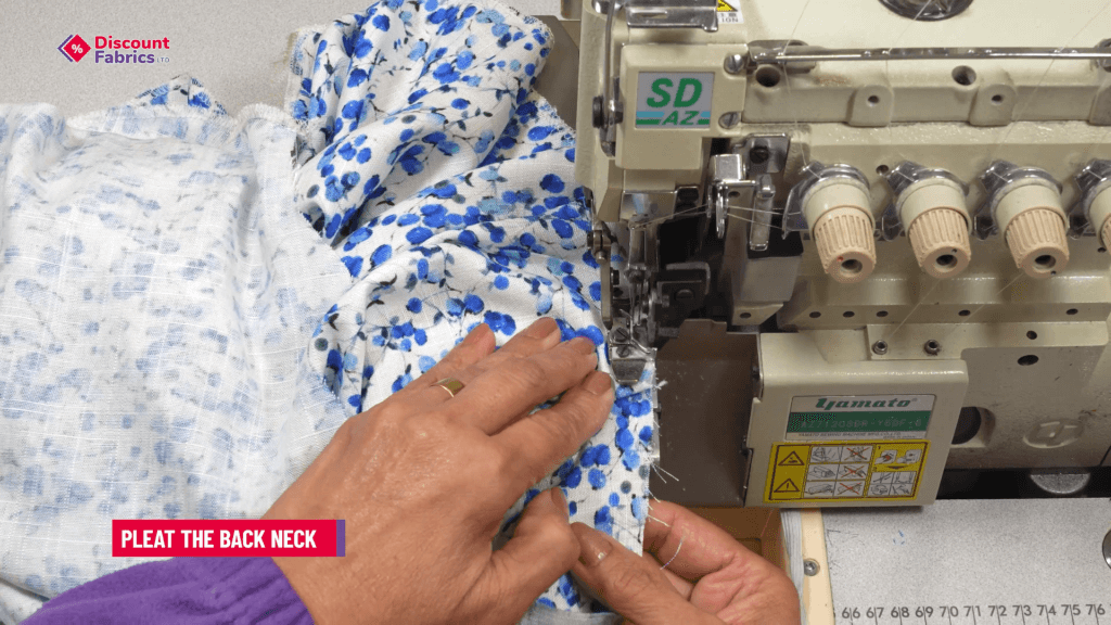 Hands pleating the back neck of a blue and white floral fabric using a Yamato sewing machine. The person is wearing a purple garment.