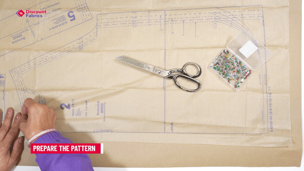 A person's hand is holding a folded paper pattern on a table. A pair of silver scissors and a plastic box of colorful pins are nearby. The text