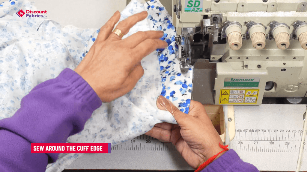 Person sewing a blue and white floral fabric with a serger, wearing a purple sweater. A label reads