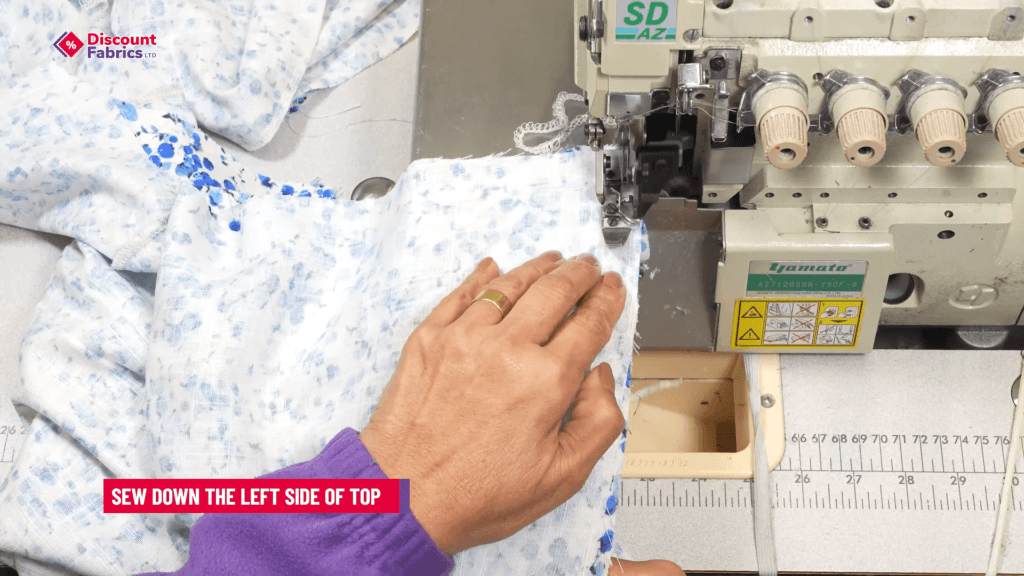 A person in a purple shirt uses a sewing machine to stitch a white fabric with blue spots. The machine has multiple knobs and spools. A red label on the image reads,